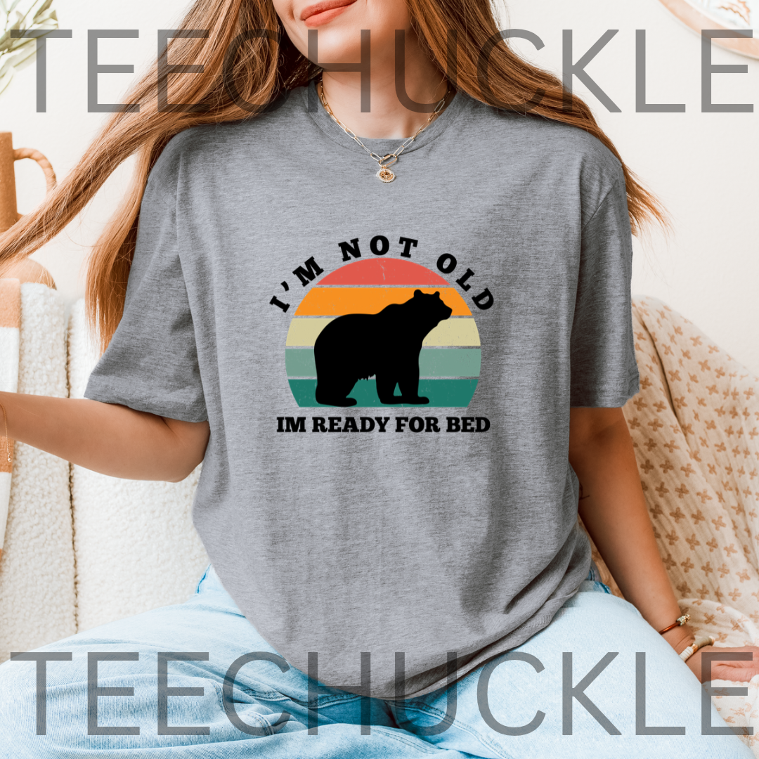 A woman wearing a gray graphic t-shirt with a retro sunset and bear design that reads 'I'M NOT OLD IM READY FOR BED'.