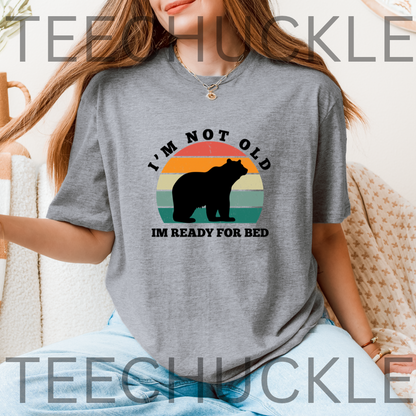 A woman wearing a gray graphic t-shirt with a retro sunset and bear design that reads 'I'M NOT OLD IM READY FOR BED'.