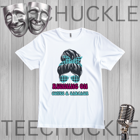 A white short-sleeve graphic t-shirt displayed on a gray wood-plank background, featuring a cartoon illustration of a woman with a plaid hair bow and sunglasses, with the text 'RUNNING ON COFFEE & SARCASM'. The image includes decorative props like two silver theater masks and a vintage-style microphone