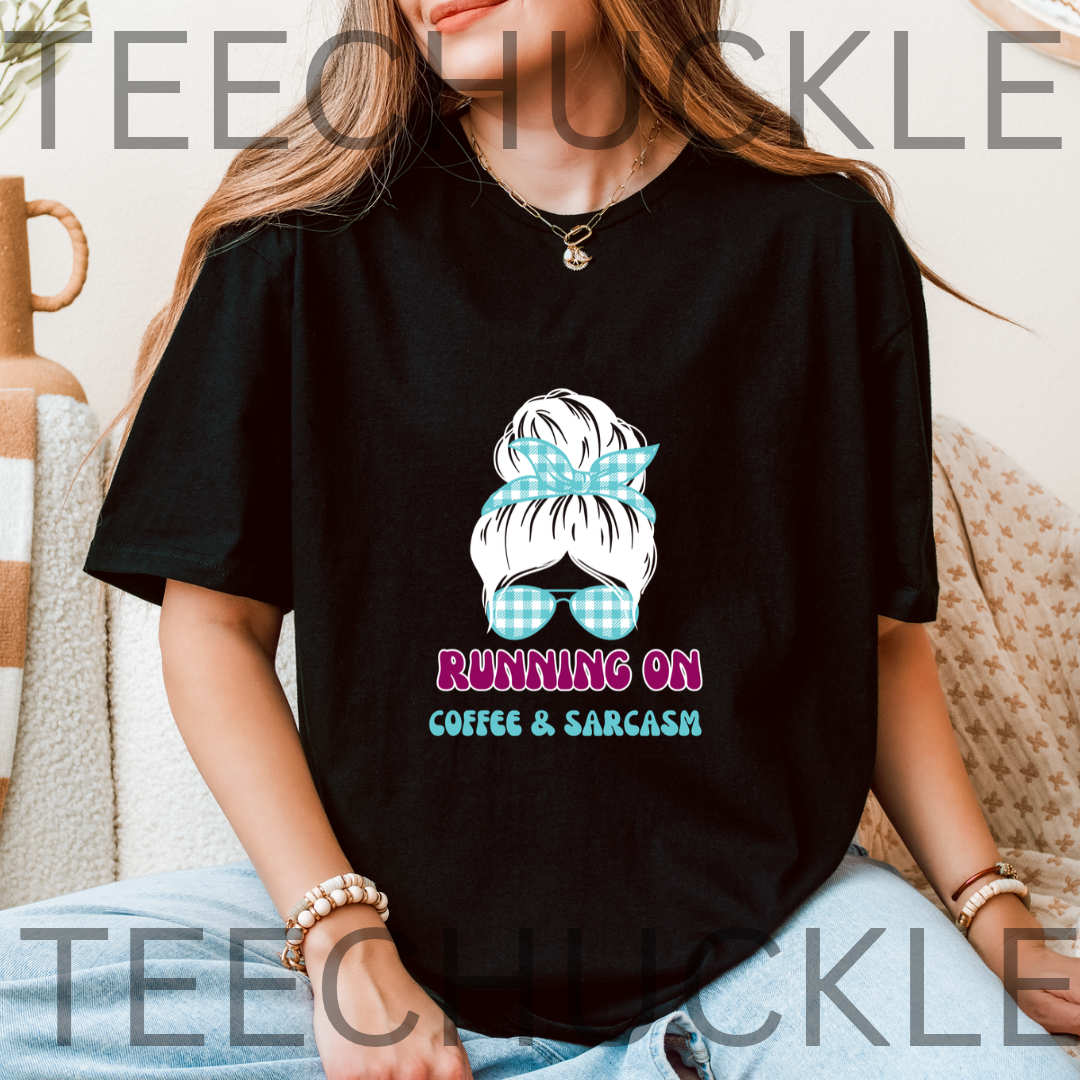 A woman wearing a black graphic t-shirt with a cartoon illustration of a woman's hair in a blue gingham bow and sunglasses, with the text 'RUNNING ON COFFEE & SARCASM' below. 