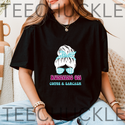 A woman wearing a black graphic t-shirt with a cartoon illustration of a woman's hair in a blue gingham bow and sunglasses, with the text 'RUNNING ON COFFEE & SARCASM' below. 