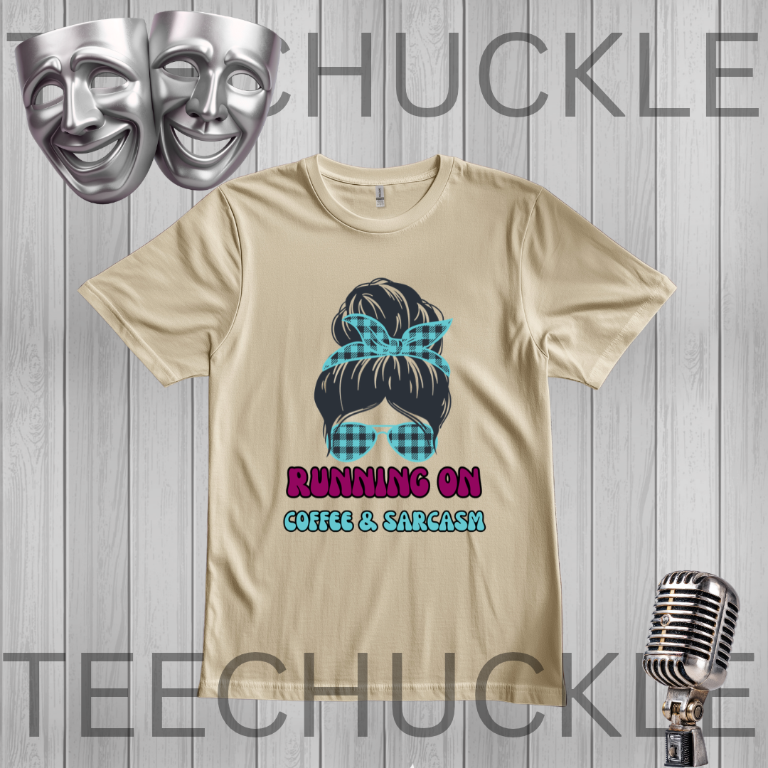 A sand graphic t-shirt featuring a cartoon illustration of a woman with a blue plaid bow and sunglasses, with the text 'RUNNING ON COFFEE & SARCASM' below. The t-shirt is displayed on a gray wood-plank background with decorative props including two silver comedy masks at the top left and a vintage silver microphone at the bottom right.