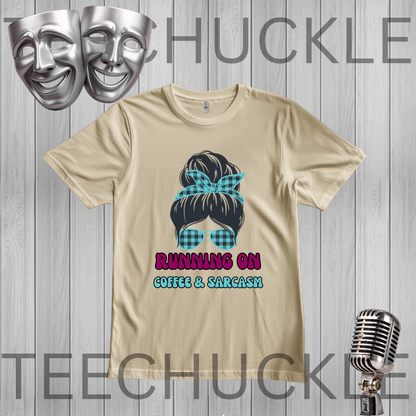 A sand graphic t-shirt featuring a cartoon illustration of a woman with a blue plaid bow and sunglasses, with the text 'RUNNING ON COFFEE & SARCASM' below. The t-shirt is displayed on a gray wood-plank background with decorative props including two silver comedy masks at the top left and a vintage silver microphone at the bottom right.