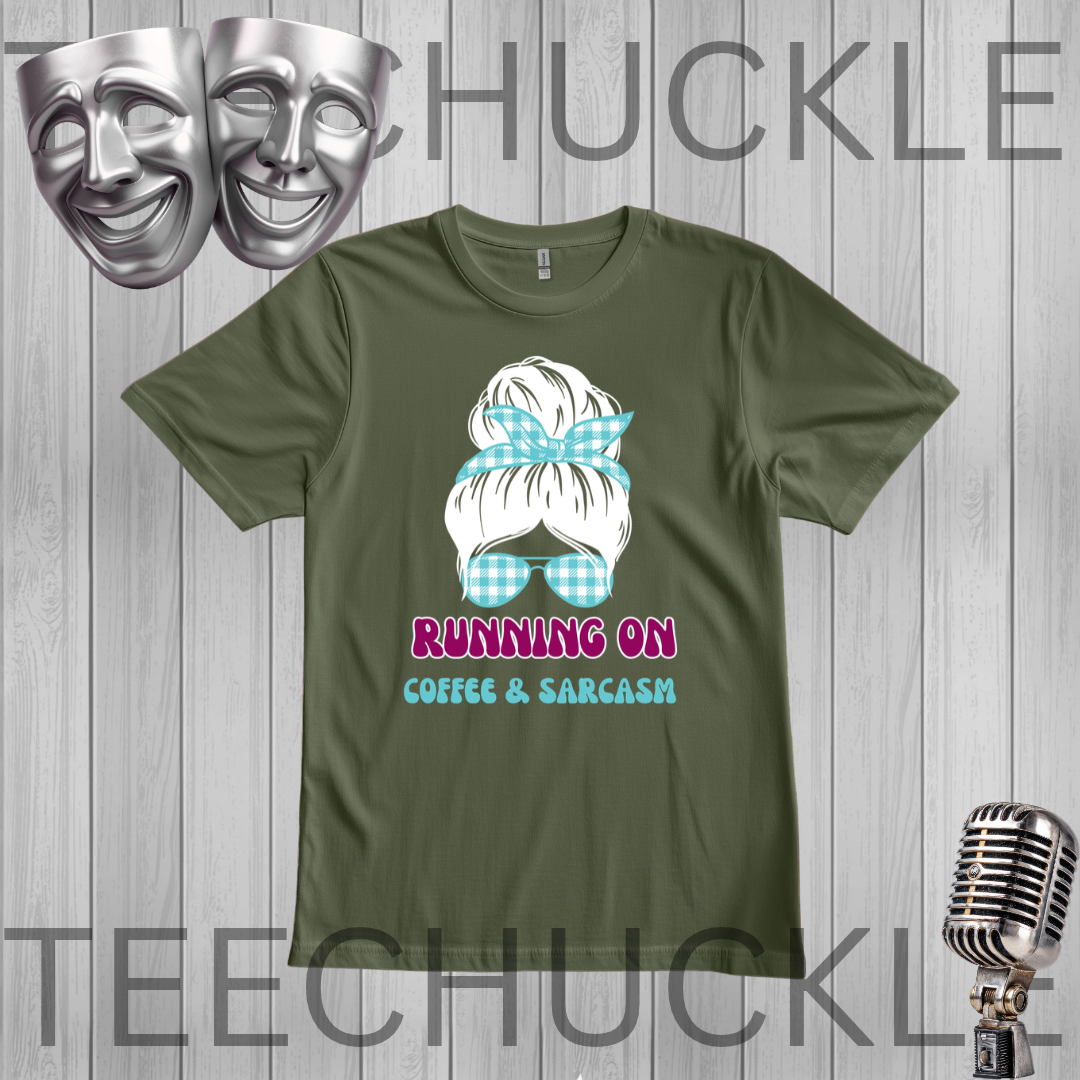 An olive green graphic t-shirt displayed on a gray wood-plank background. The graphic features a white hair bun with a blue and white gingham bow and matching gingham glasses, with the text 'RUNNING ON COFFEE & SARCASM' in pink and light blue.