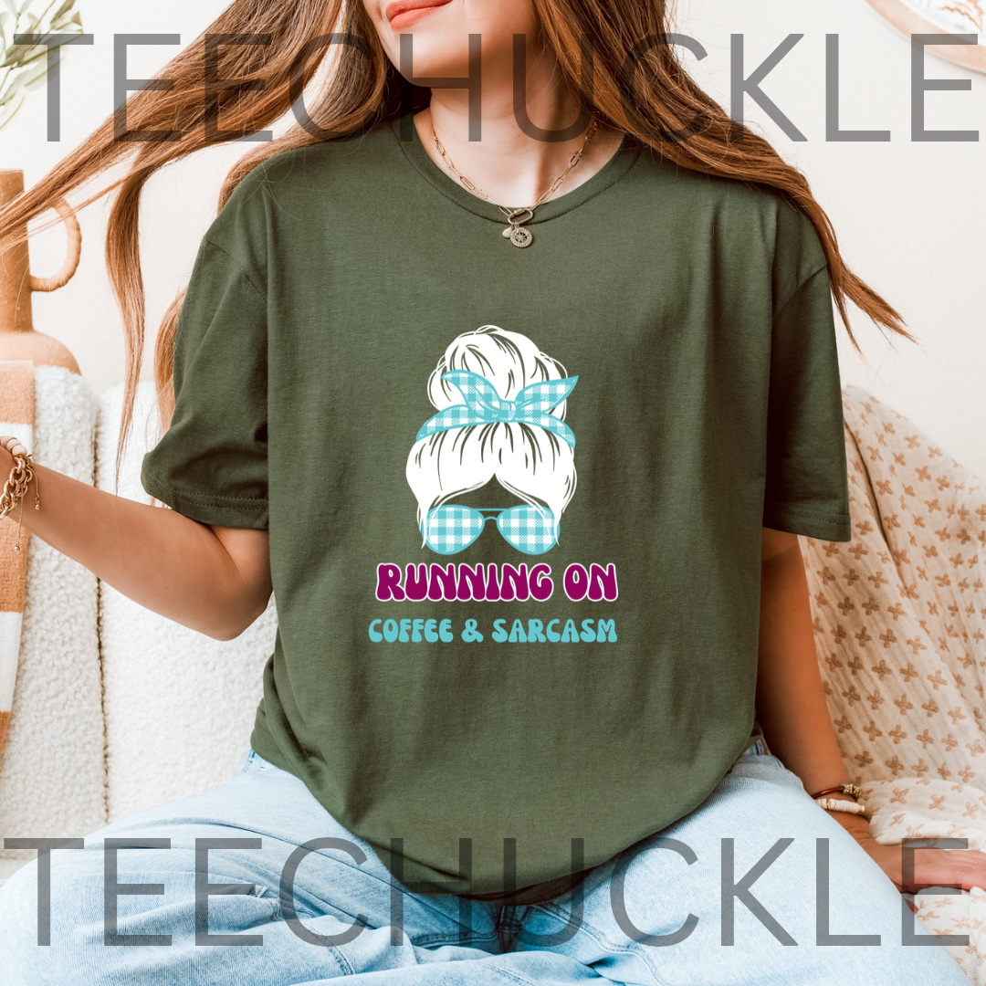 A woman wearing a green graphic t-shirt with a cartoon illustration of a woman's hair in a messy bun with a blue gingham bow and the text 'RUNNING ON COFFEE & SARCASM'.