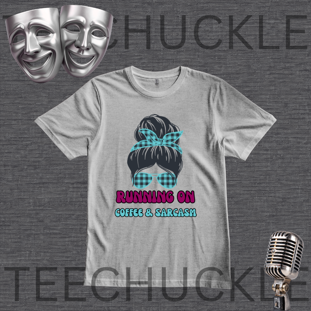 A gray graphic t-shirt featuring a black hair bun with a teal plaid bow and matching plaid sunglasses, with the text 'RUNNING ON COFFEE & SARCASM' in pink and teal. The t-shirt is displayed on a dark gray textured background with theatrical masks, a vintage microphone, and a watermark.