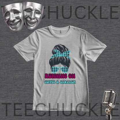 A gray graphic t-shirt featuring a black hair bun with a teal plaid bow and matching plaid sunglasses, with the text 'RUNNING ON COFFEE & SARCASM' in pink and teal. The t-shirt is displayed on a dark gray textured background with theatrical masks, a vintage microphone, and a watermark.