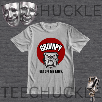 A gray graphic t-shirt featuring a red circle with the word 'GRUMPY' above a bulldog illustration and the phrase 'GET OFF MY LAWN.' The shirt is displayed on a dark gray textured background with theatrical comedy masks in the top-left and a vintage microphone in the bottom-right.
