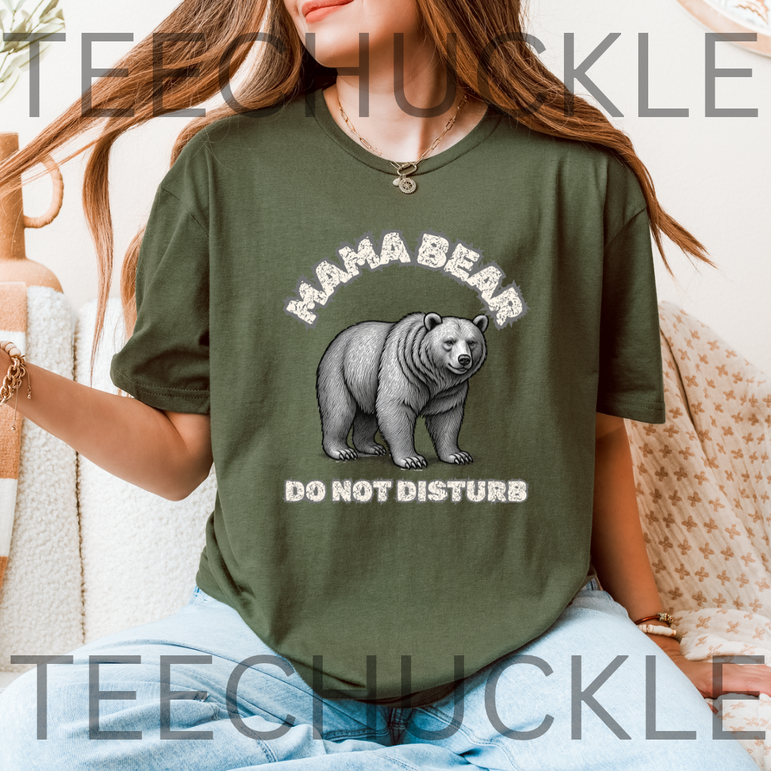A woman wearing a green graphic t-shirt with a 'MAMA BEAR DO NOT DISTURB' print featuring a bear illustration, set against a cozy indoor background with a blanket.