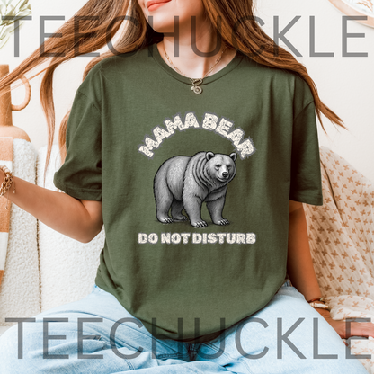 A woman wearing a green graphic t-shirt with a 'MAMA BEAR DO NOT DISTURB' print featuring a bear illustration, set against a cozy indoor background with a blanket.