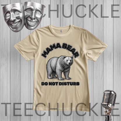 A sand graphic t-shirt featuring a black and white illustration of a mama bear with the text 'MAMA BEAR DO NOT DISTURB'. The t-shirt is displayed on a gray wood-plank background with decorative props including two silver theater masks, a vintage microphone