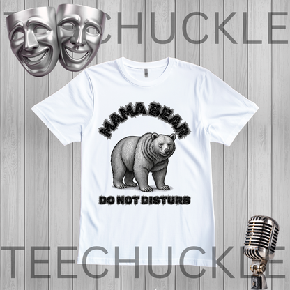 A white graphic t-shirt featuring a black and gray illustration of a bear with the text 'MAMA BEAR DO NOT DISTURB'. The t-shirt is displayed on a gray wood-plank background with decorative props including two silver theater masks at the top left, a vintage silver microphone at the bottom right