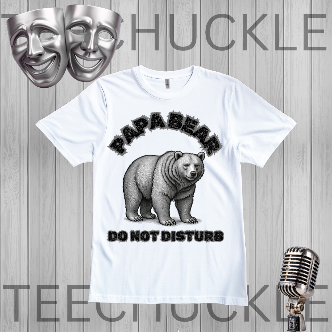 A white graphic t-shirt featuring a black and gray illustration of a bear with the text 'PAPABEAR DO NOT DISTURB'. The t-shirt is displayed on a gray wood-plank background with decorative props including two silver theater masks at the top left, a vintage-style microphone at the bottom right