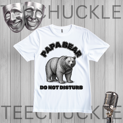 A white graphic t-shirt featuring a black and gray illustration of a bear with the text 'PAPABEAR DO NOT DISTURB'. The t-shirt is displayed on a gray wood-plank background with decorative props including two silver theater masks at the top left, a vintage-style microphone at the bottom right