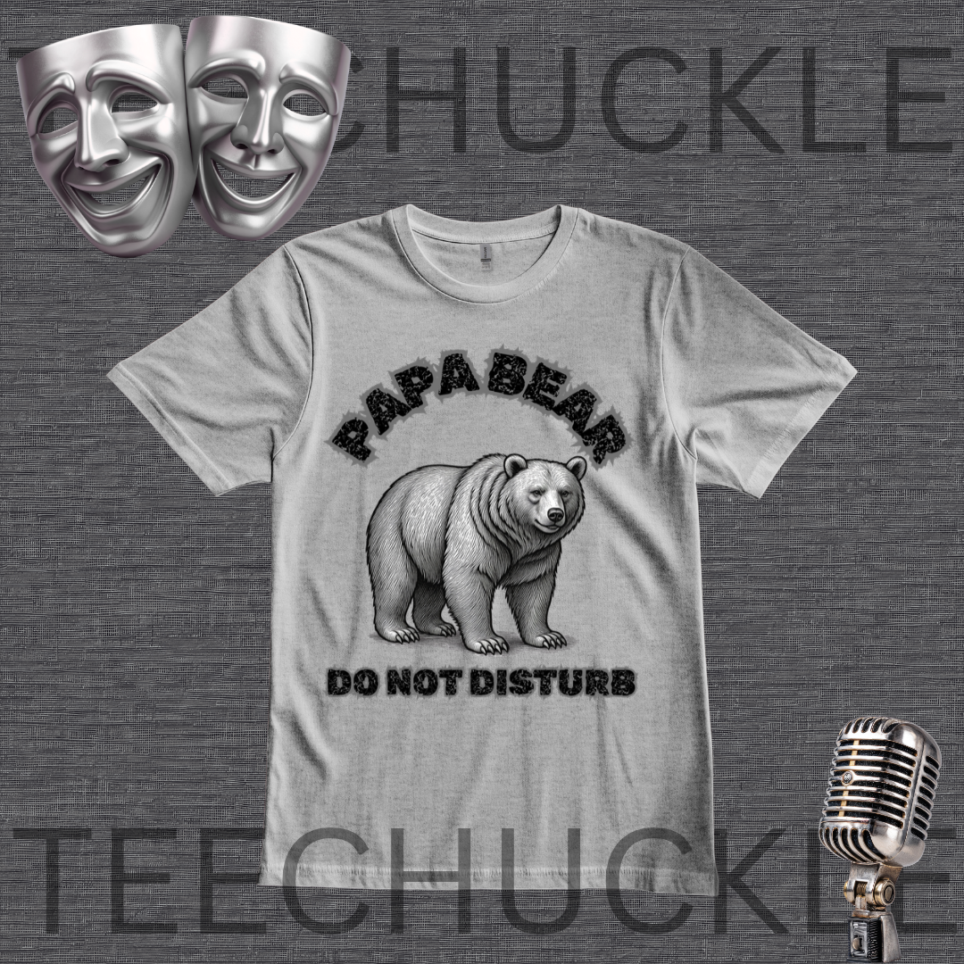 A gray graphic t-shirt featuring a black and white illustration of a bear with the text 'PAPA BEAR DO NOT DISTURB'. The t-shirt is displayed on a dark gray textured background with decorative props including two silver comedy masks, a vintage microphone,