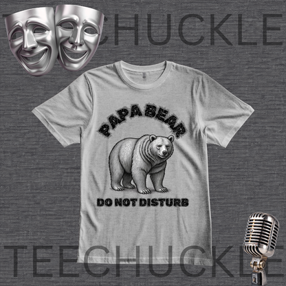 A gray graphic t-shirt featuring a black and white illustration of a bear with the text 'PAPA BEAR DO NOT DISTURB'. The t-shirt is displayed on a dark gray textured background with decorative props including two silver comedy masks, a vintage microphone,