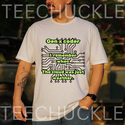 A man wearing a white graphic t-shirt with a green and black circuit board design and the text 'Gen x coder I remember when The cloud was just weather.' The background is a blurred outdoor setting