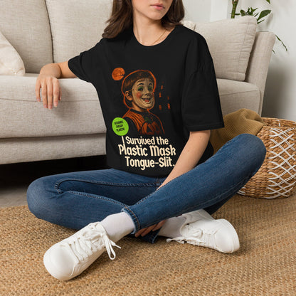 A woman sits cross-legged on a woven rug in front of a beige sofa, wearing a black graphic T-shirt with a cchild in halloween mask illustration and the text 'I Survived the Plastic Mask Tongue-Slit.' She is also wearing blue jeans and white sneakers.