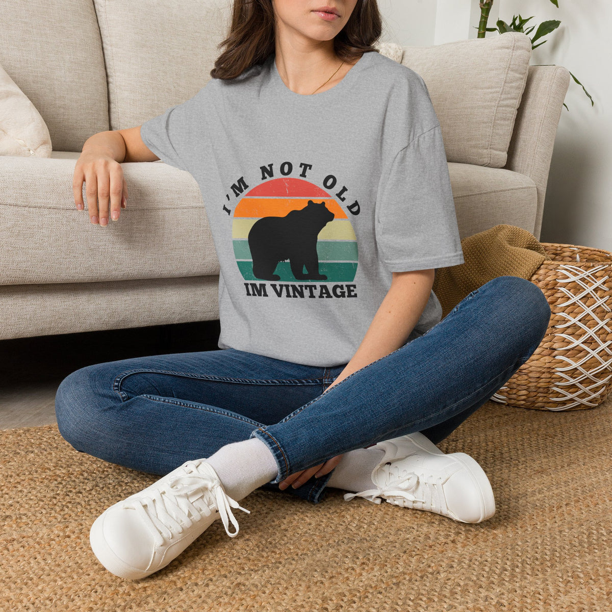 A woman sitting cross-legged on a woven rug in front of a beige sofa, wearing a gray graphic T-shirt with a bear silhouette and retro sunset design reading 'I'M NOT OLD I'M VINTAGE,' paired with blue jeans and white sneakers.