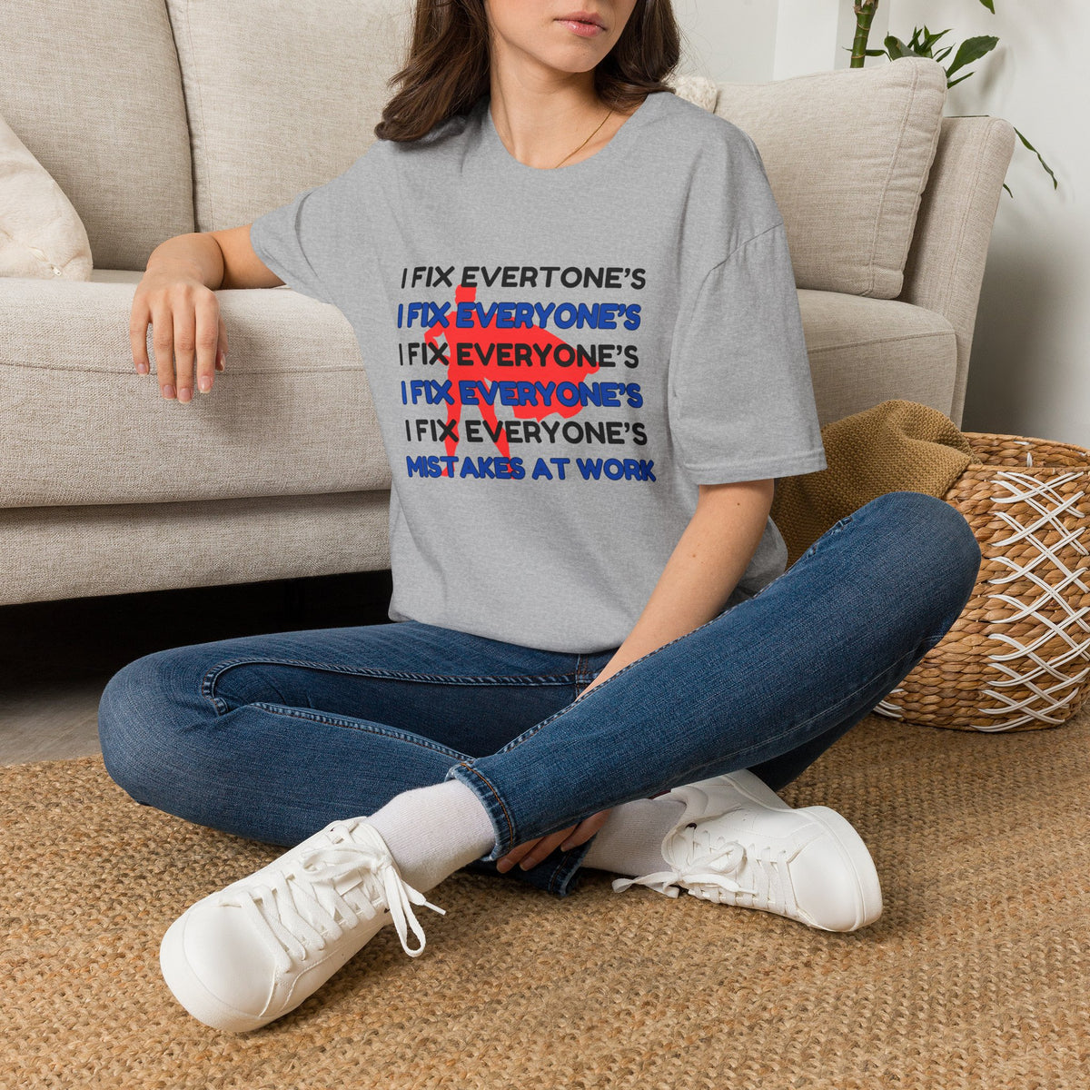A woman sits cross-legged on a woven rug in a living room, wearing a light gray graphic T-shirt with the phrase 'I FIX EVERYONE'S MISTAKES AT WORK' repeated in black, blue, and red, paired with blue jeans and white sneakers.