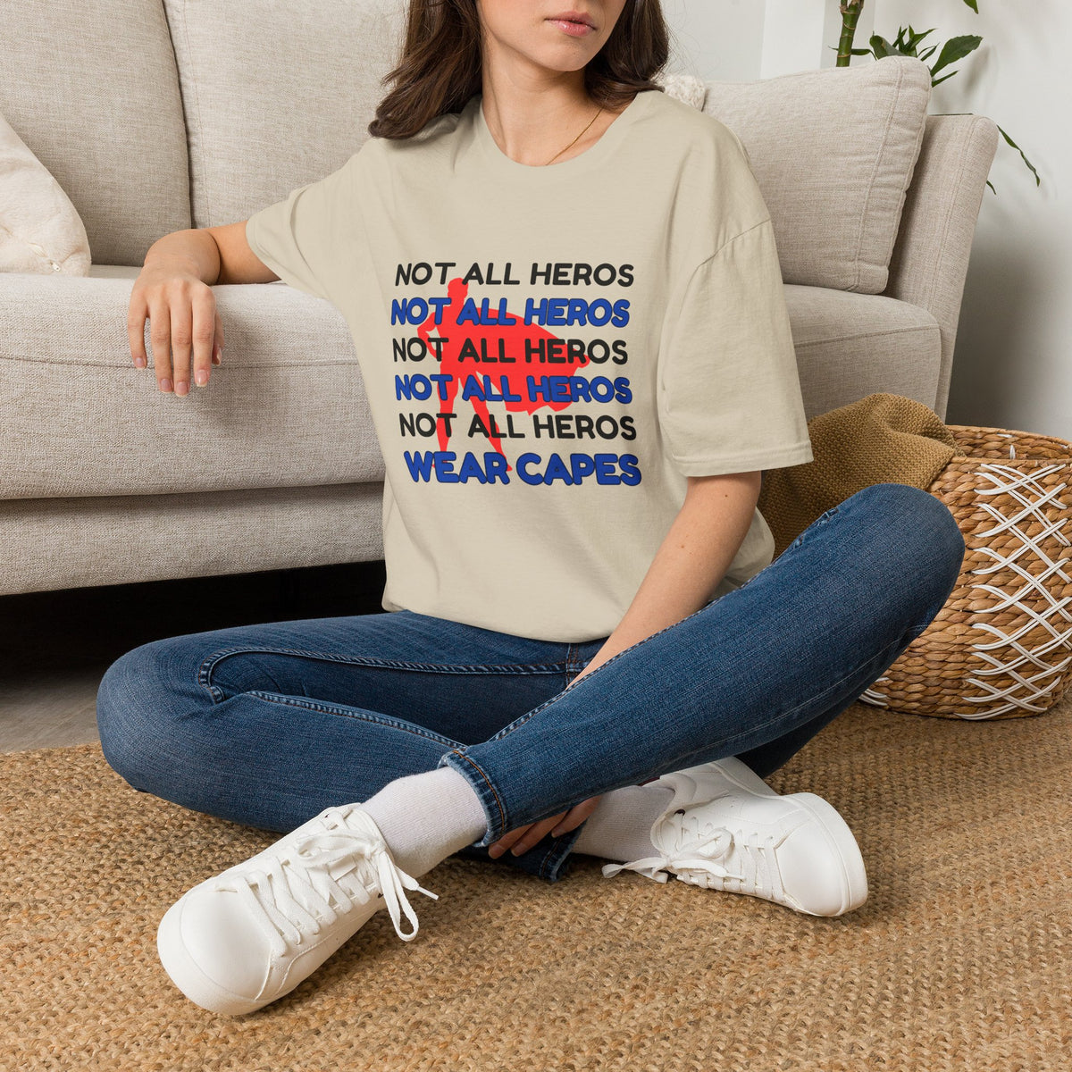 A woman sitting cross-legged on a woven rug in front of a beige sofa, wearing a sand graphic T-shirt with the text 'NOT ALL HEROS' repeated in black, blue, and red superhero behinf it, with 'WEAR CAPES' in blue at the bottom.