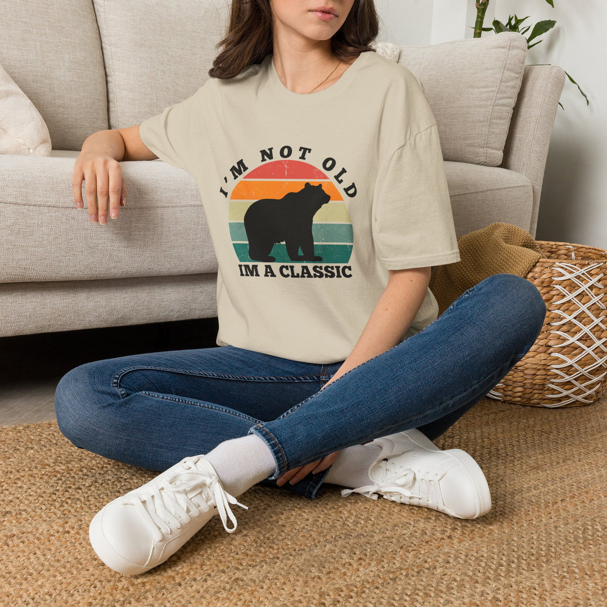 A woman sitting cross-legged on a woven rug in front of a light beige sofa, wearing a sand graphic T-shirt with a bear silhouette and the text 'I'M NOT OLD I'M A CLASSIC' over a retro sunset design, paired with blue jeans and white sneakers.