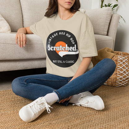 A woman sitting cross-legged on a woven rug in front of a beige sofa, wearing a beige graphic T-shirt with a black circular logo that reads 'Vintage Bos Hip Hop Scratched But Still A Classic' and blue jeans with white sneakers.