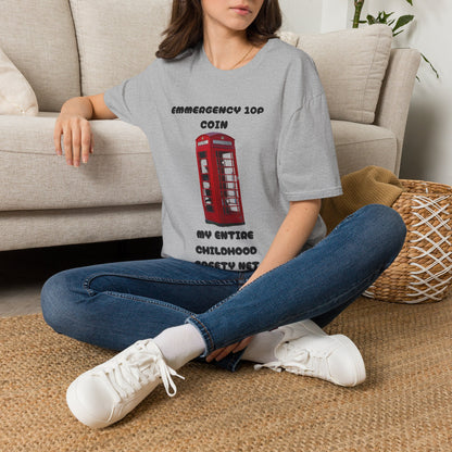 A woman sitting cross-legged on a woven rug in front of a beige sofa, wearing a light graphic T-shirt with a red telephone booth print and the text 'EMMERGENCY 10P COIN MY ENTIRE CHILDHOOD SAFETY NET', paired with blue jeans and white sneakers.