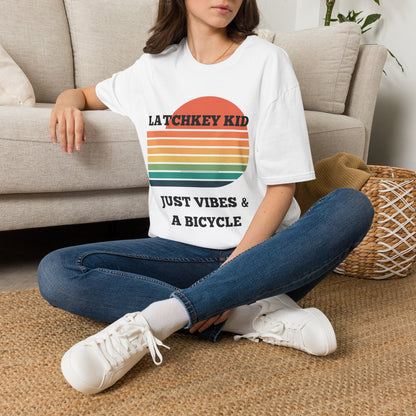 A woman sitting cross-legged on a woven rug in front of a beige couch, wearing a white graphic T-shirt with a retro sunset design and the text 'LATCHKEY KID JUST VIBES & A BICYCLE'.
