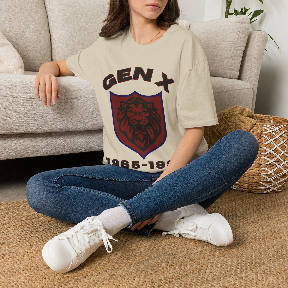 A woman sitting cross-legged on a woven rug in front of a light gray sofa, wearing a sand graphic T-shirt with a dark red lion crest and the text 'GEN-X 1965-1991', blue jeans, and white sneakers.