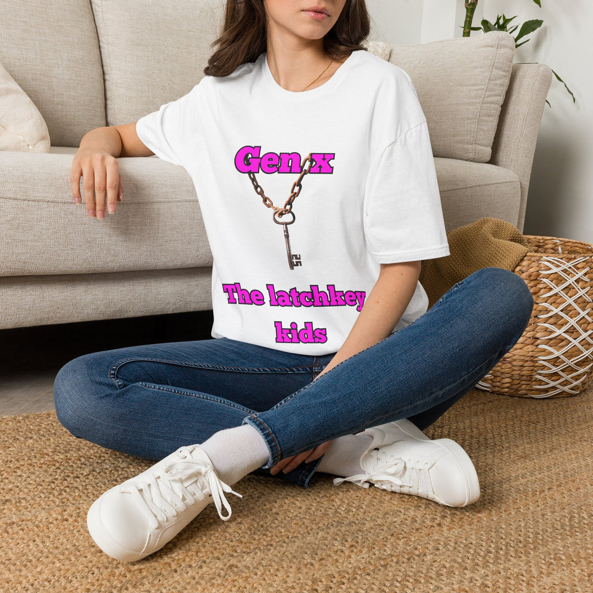 A woman sitting cross-legged on a woven rug in front of a beige sofa, wearing a white graphic T-shirt with pink text reading 'Gen x The latchkey kids' and a key-and-chain design, paired with blue jeans and white sneakers.