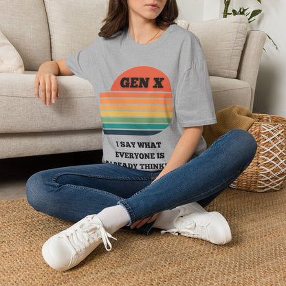 A woman sitting cross-legged on a woven rug in front of a beige sofa, wearing a gray graphic T-shirt with a retro sunset design and the text 'GEN X I SAY WHAT EVERYONE IS ALREADY THINKING'.