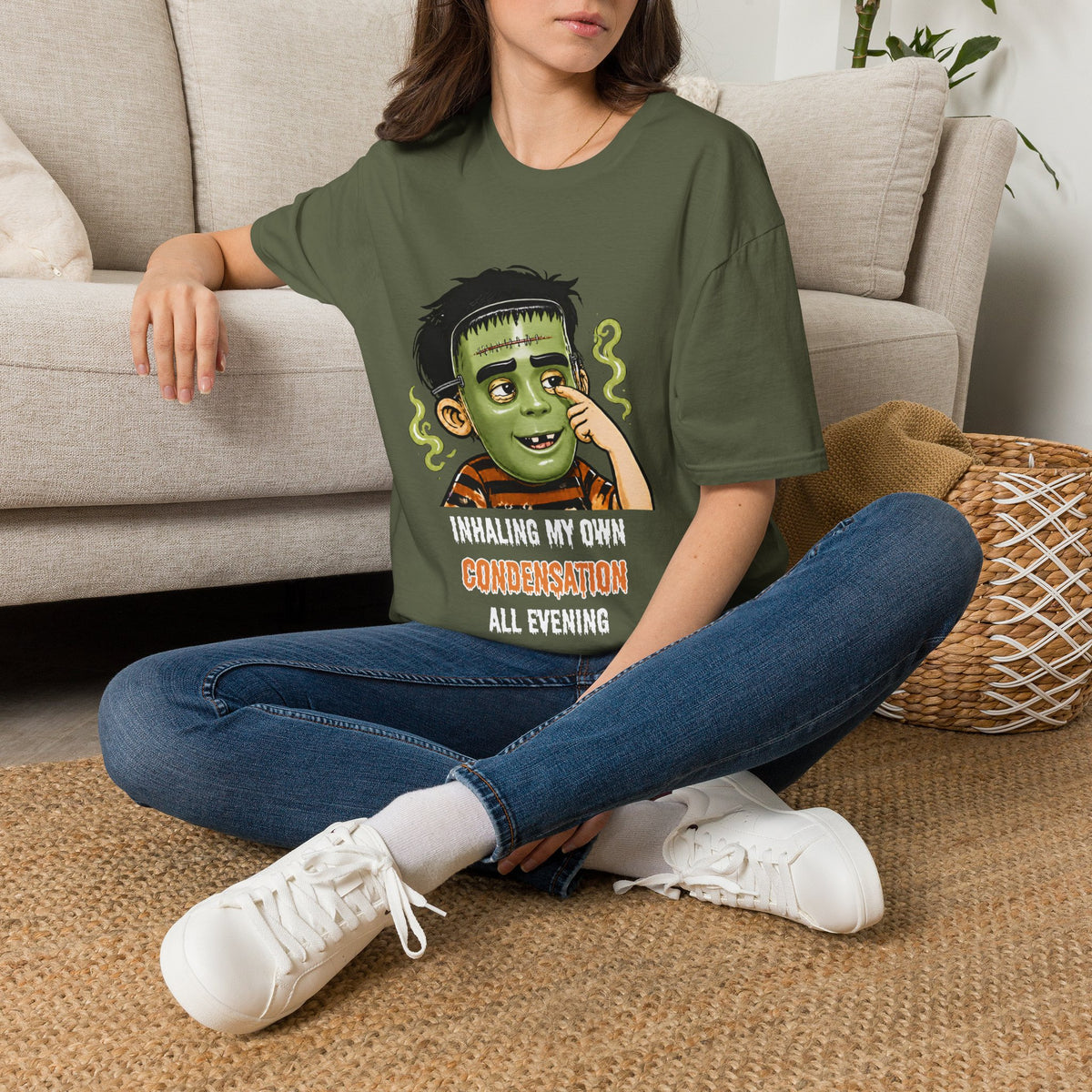 A woman sitting on a woven rug in front of a beige sofa, wearing an olive green graphic t-shirt with a child in a halloween mask print that reads 'INHALE MY OWN CONDENSATION ALL EVENING'.