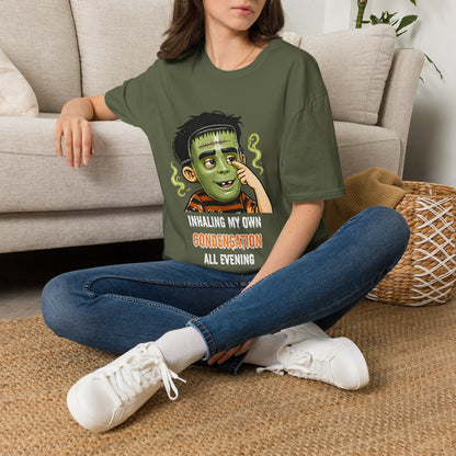 A woman sitting on a woven rug in front of a beige sofa, wearing an olive green graphic t-shirt with a child in a halloween mask print that reads 'INHALE MY OWN CONDENSATION ALL EVENING'.