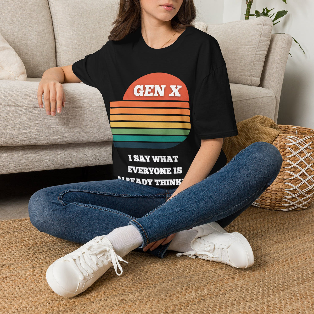 A woman sitting on a woven rug in front of a beige sofa, wearing a black graphic T-shirt with a retro sunset design and the text 'GEN X I SAY WHAT EVERYONE IS ALREADY THINKING'.