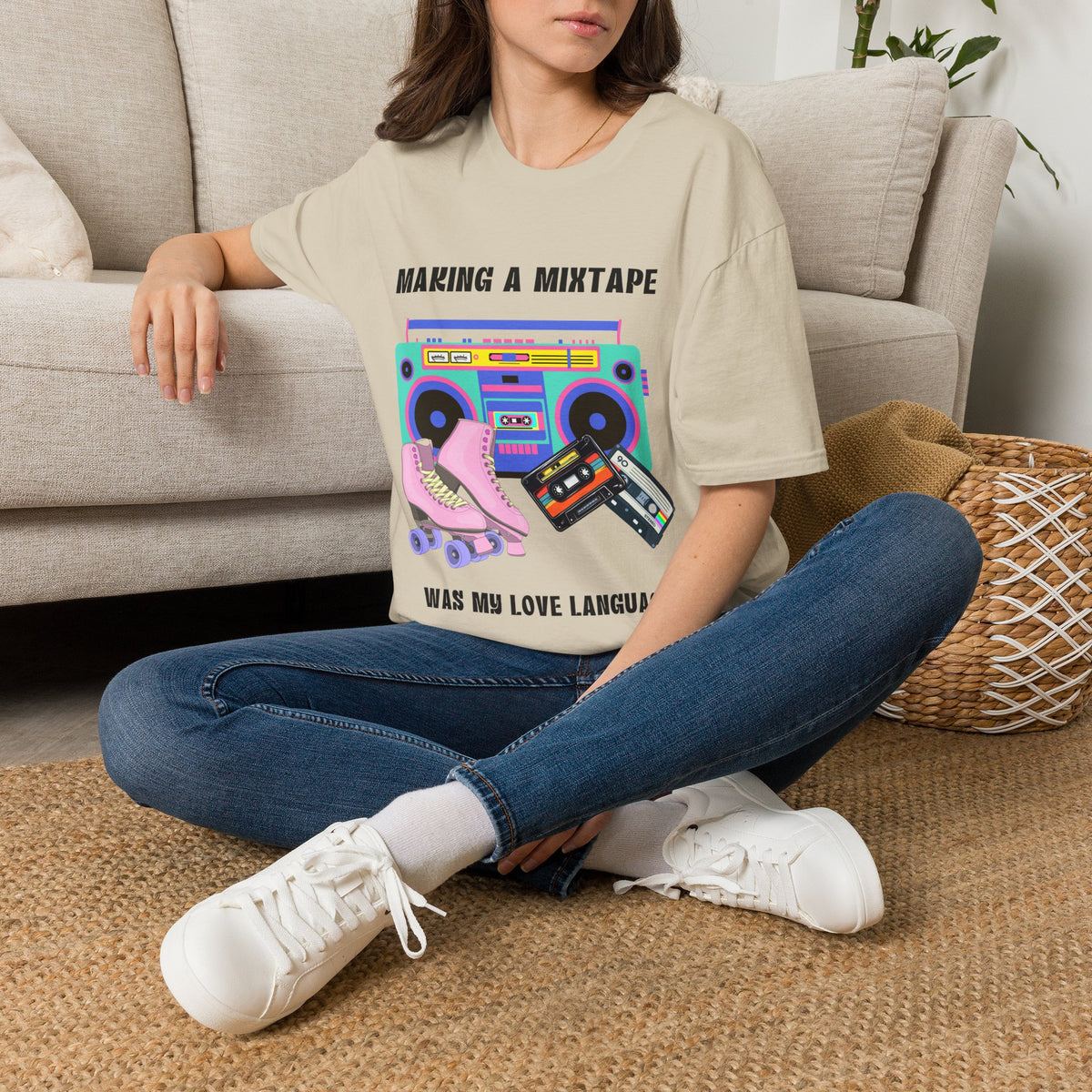 A woman sitting cross-legged on a woven rug in front of a beige sofa, wearing a sand graphic T-shirt with a retro boombox, cassette tape, and roller skates design, with the text "making a mixtape was my love language" with blue jeans, and white sneakers.