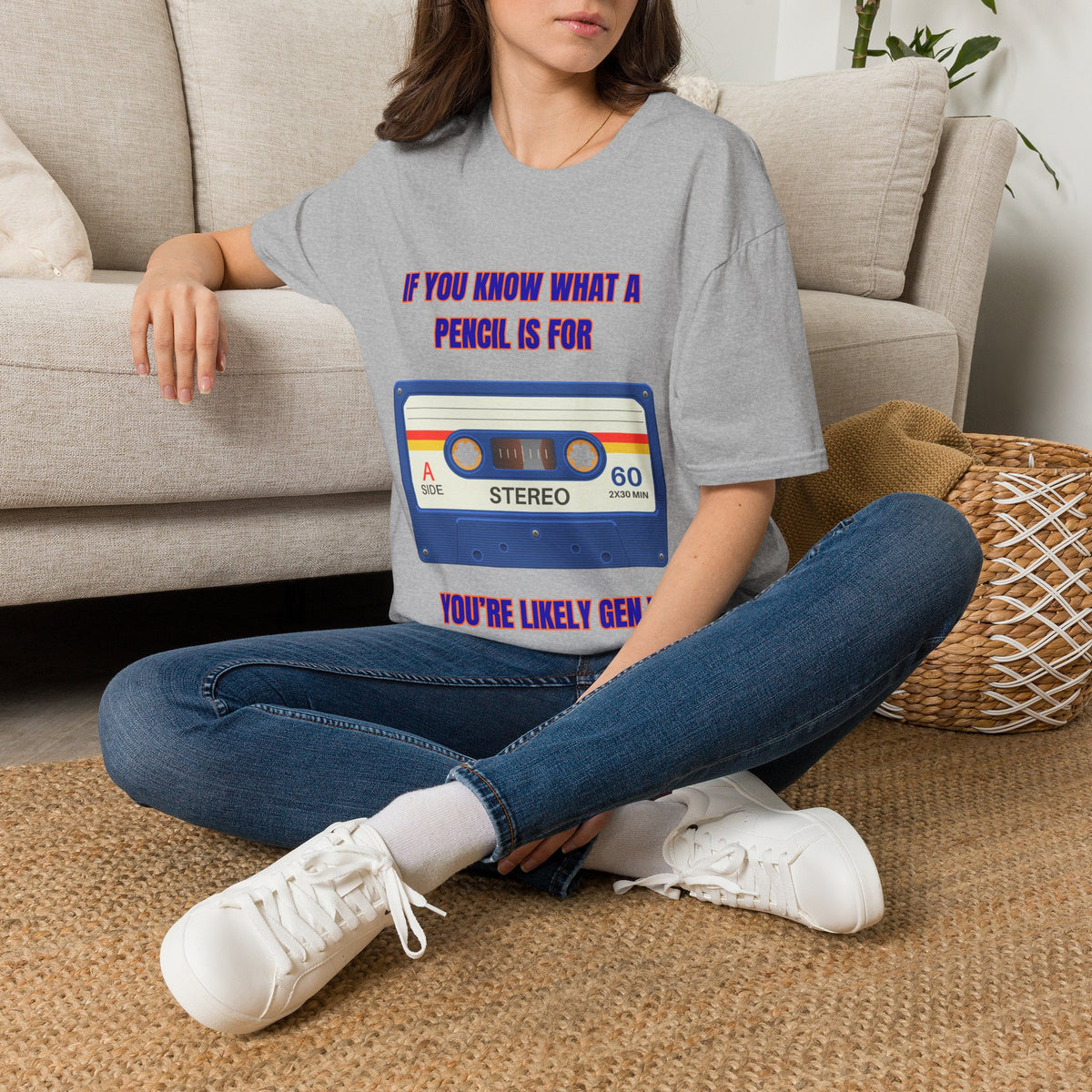 A woman sitting on a woven rug in front of a beige sofa, wearing a gray graphic t-shirt with a retro cassette tape design and text " IF YOU KNOW WHAT A PENCIL IS FOR YOU'RE LIKELY GEN X" and blue jeans.
