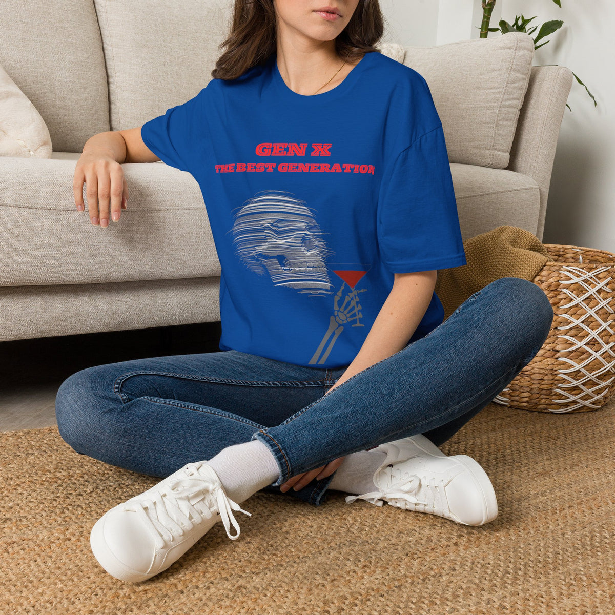 A woman sat on a rug next to a couch wearing a blue short-sleeve graphic T-shirt, featuring red text reading 'GEN X THE BEST GENERATION' above a gray illustrated figure holding a red martini glass. 