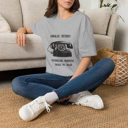 A woman sitting cross-legged on a woven rug in front of a beige sofa, wearing a  gray graphic T-shirt. The shirt has a black printed design featuring a vintage rotary telephone and the text 'ANALOGIC MEMORY' and 'MEMORIZING NUMBERS INSIDE MY HEAD. paired with blue jeans and white sneakers.