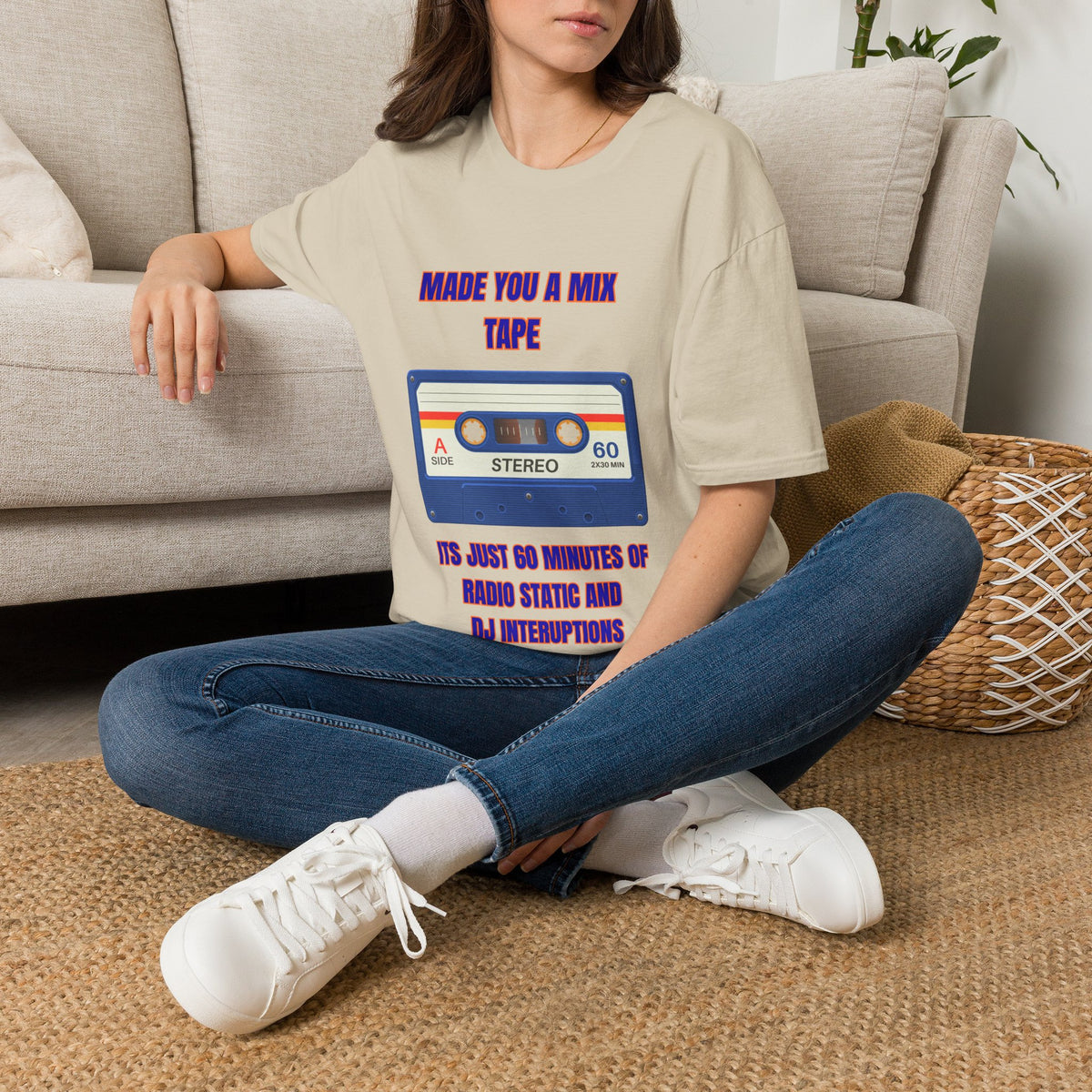 A woman sits cross-legged on a woven rug in front of a beige sofa, wearing a beige graphic T-shirt with a retro cassette tape design and text that reads 'MADE YOU A MIX TAPE' and 'ITS JUST 60 MINUTES OF RADIO STATIC AND DJ INTERRUPTIONS.' She is also wearing blue jeans and white sneakers.
