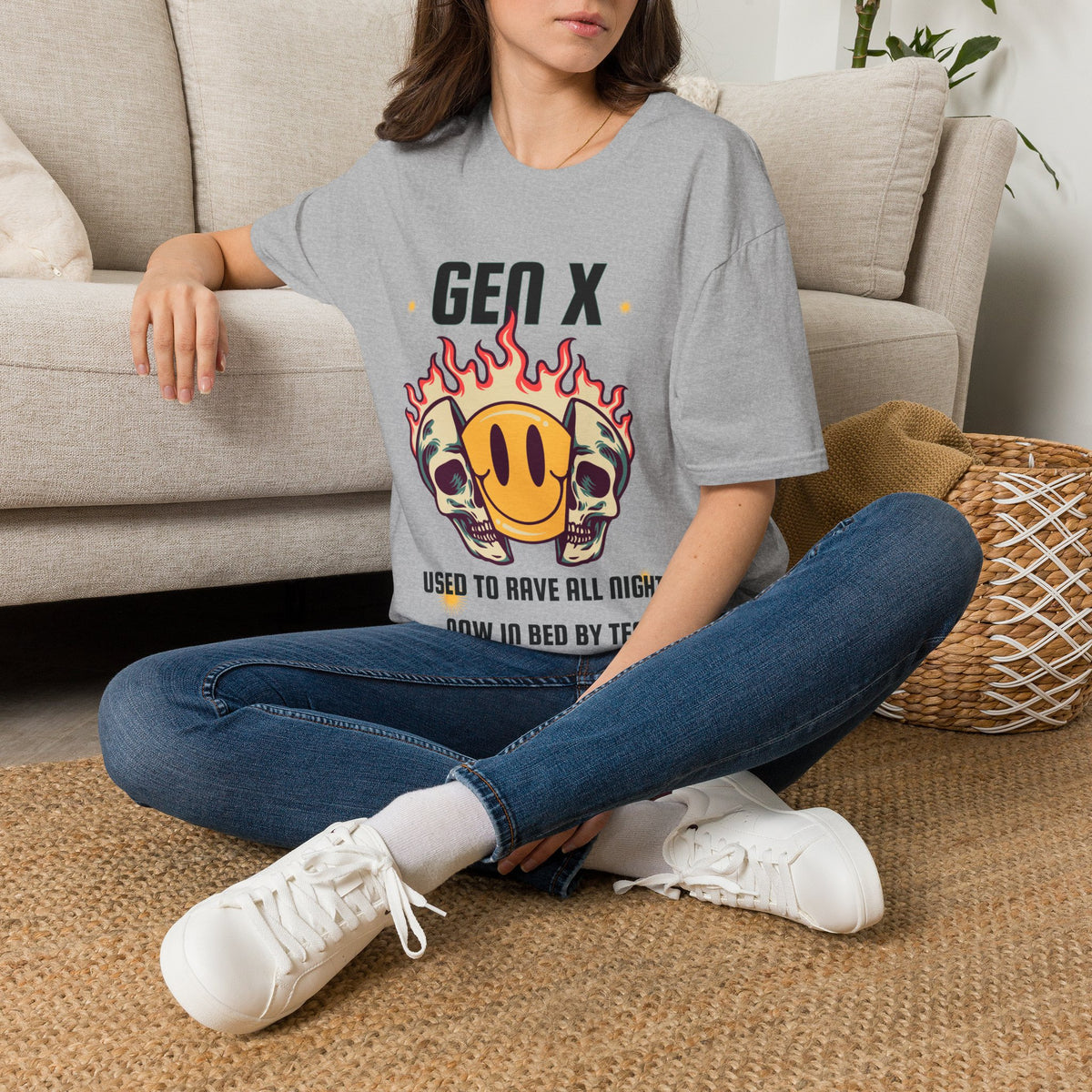A woman sitting cross-legged on a woven rug in front of a beige sofa, wearing a lgray graphic T-shirt with a skull-and-smiley-face design 