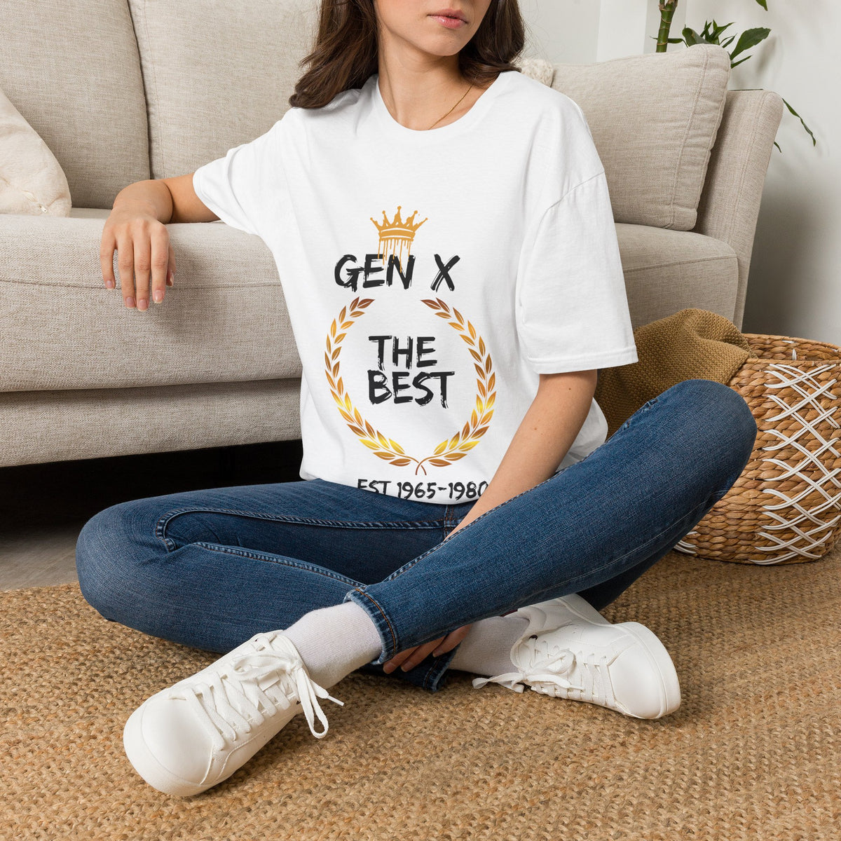 Person sitting on the floor wearing a white t-shirt with a Gen X The Best graphic design with crown and gold wreath design, wearing blue jeans, and white sneakers.