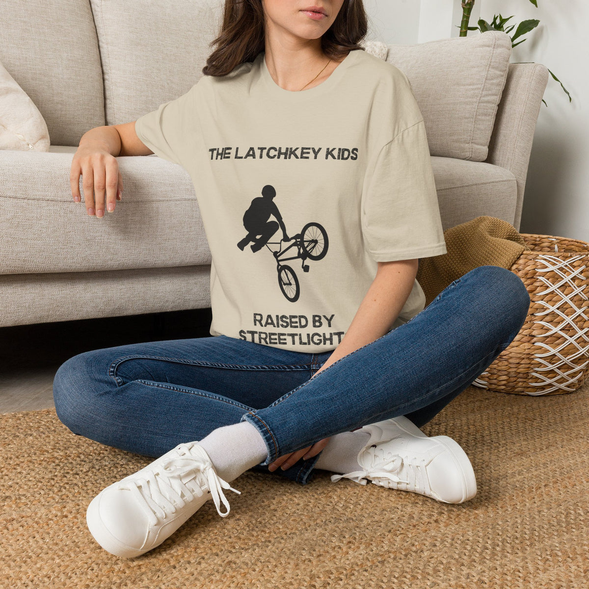A woman sits cross-legged on a woven rug in front of a beige sofa, wearing an oversized sand graphic T-shirt with black text reading 'THE LATCHKEY KIDS' and 'RAISED BY STREETLIGHT' around a BMX rider graphic, paired with blue jeans and white sneakers.