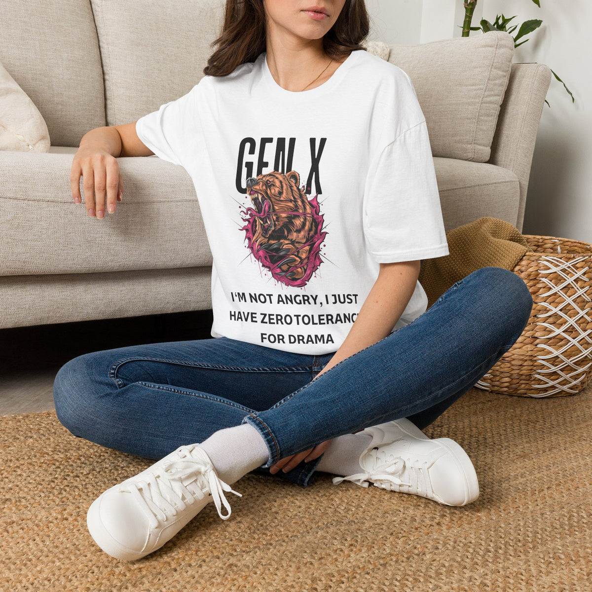 A woman sitting cross-legged on a woven rug in front of a beige sofa, wearing a white graphic T-shirt with a roaring bear illustration with pink and black details, accompanied by the text 'GEN X' and 'PROTECTIVE BY NATURE, CYNICAL BY CHOICE'. and blue jeans with white sneakers.