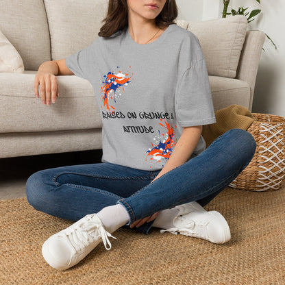 A woman sitting cross-legged on a woven rug in front of a beige sofa, wearing a gray graphic T-shirt with orange, blue, and black abstract designs and the text 'RAISED ON GRUNGE & ATTITUDE'.