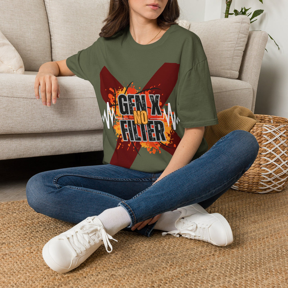 A woman sits cross-legged on a woven rug in front of a light beige sofa, wearing a green graphic T-shirt with a large red 'X' and the text 'GEN X NO FILTER'. She is also wearing blue jeans and white sneakers.