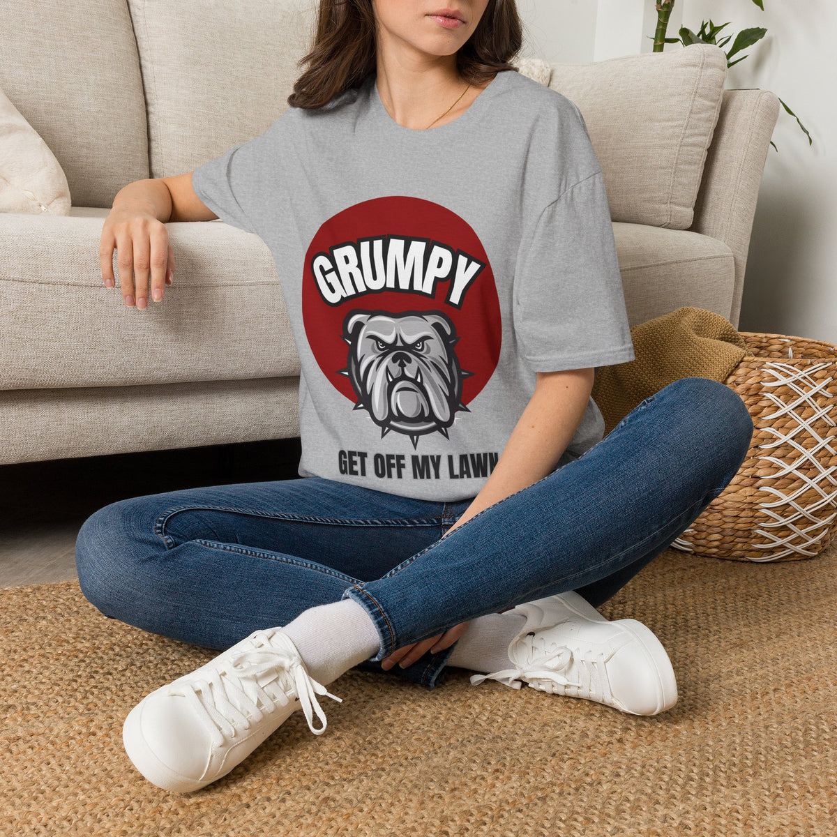 A woman sitting on a woven rug in front of a beige sofa, wearing a gray graphic t-shirt with a red circle and bulldog design that reads 'GRUMPY GET OFF MY LAWNS'.