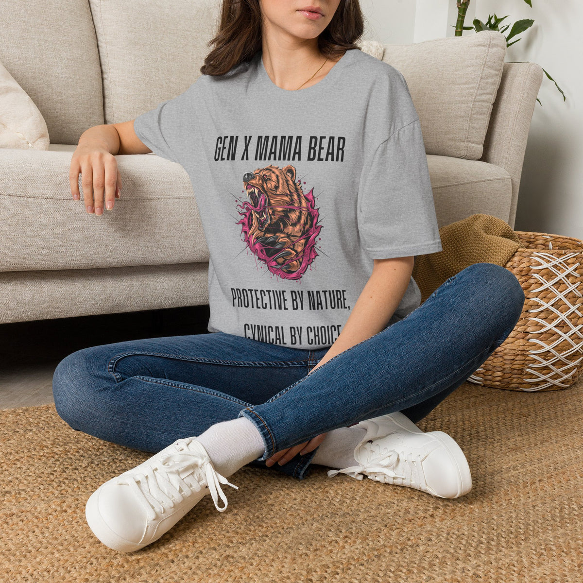 A woman sitting cross-legged on a woven rug in front of a beige sofa, wearing a gray graphic T-shirt with the text 'GEN X MAMA BEAR' and 'PROTECTIVE BY NATURE, CYGNICAL BY CHOICE' around a bear illustration, paired with blue jeans and white sneakers.