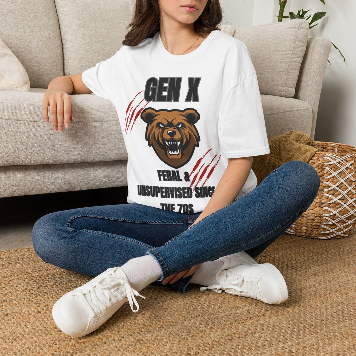 A woman sitting on a woven rug in front of a beige sofa, wearing a white graphic T-shirt with a bear illustration and the text 'GEN X FERAL & UNSUPERVISED SINCE THE 7OS'.