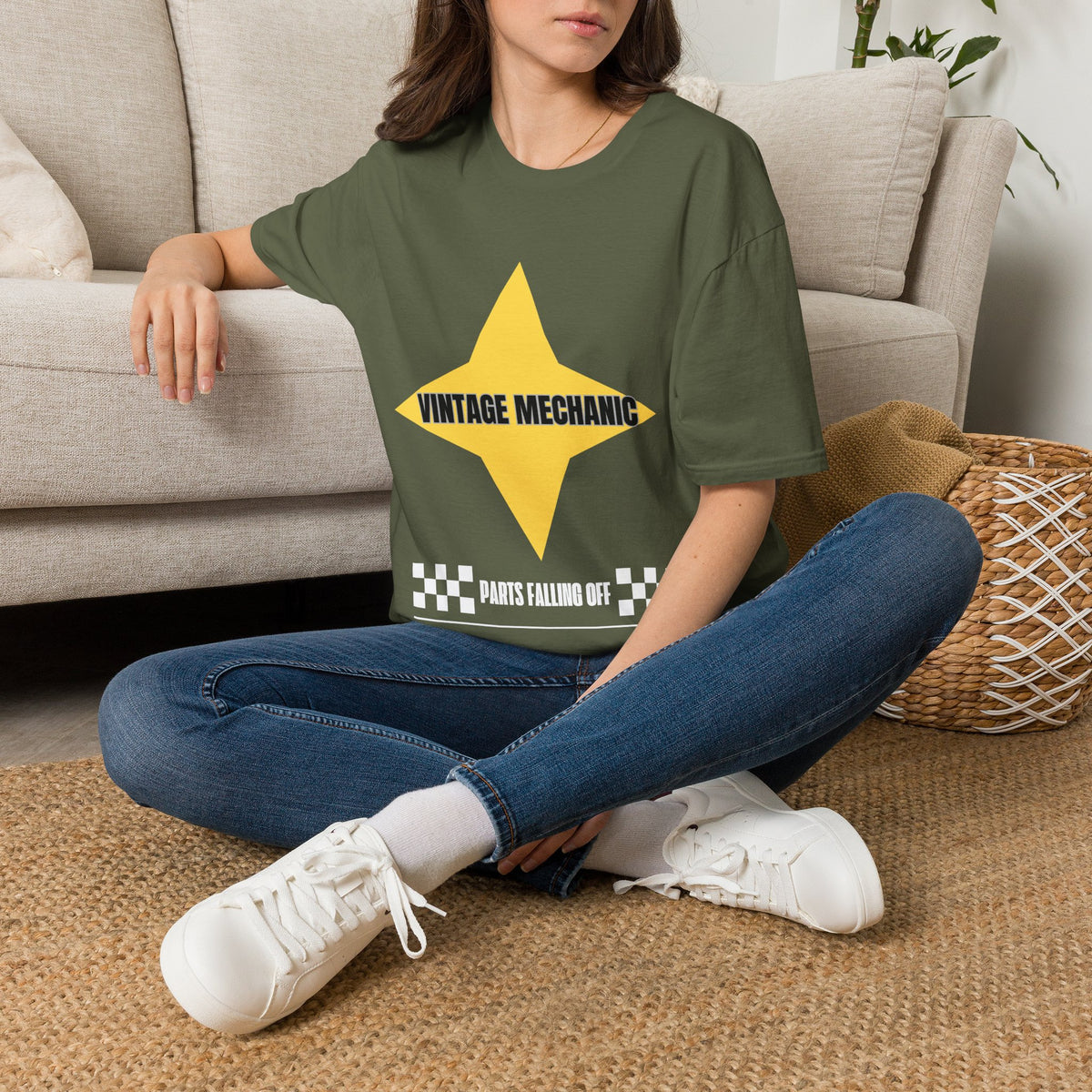 A woman sitting cross-legged on a woven rug in front of a beige sofa, wearing an green graphic T-shirt with a large yellow star and the text 'VINTAGE MECHANIC' and 'PARTS FALLING OFF'.